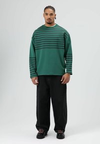 Groen shirt met lange mouwen en zwarte horizontale strepen, gecombineerd met losse zwarte spijkerbroek en zwarte schoenen met roze veters.