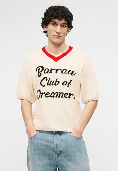 Jeune homme aux cheveux bouclés portant un pull crème avec un col rouge et le texte noir "Barrow Club of Dreamers" ainsi qu'un jean bleu clair, mains dans les poches.