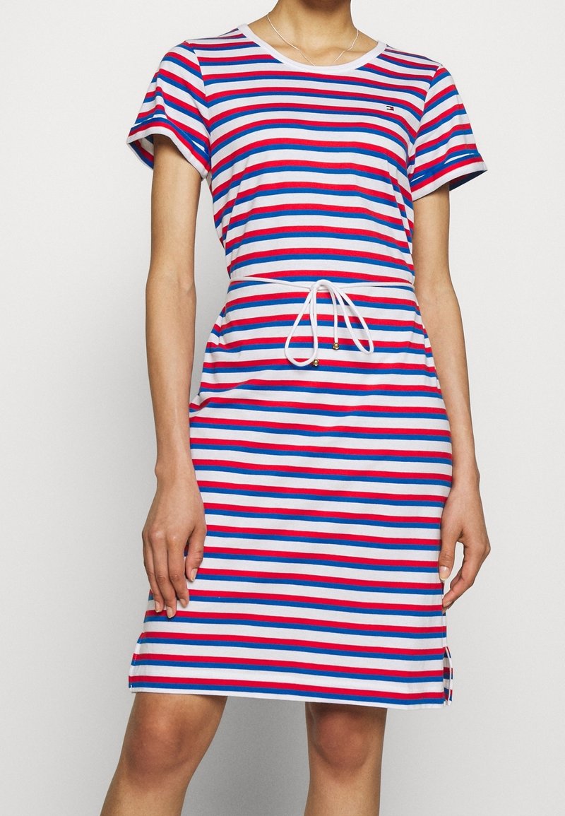 Femme portant une robe mi-longue à manches courtes avec des rayures horizontales rouges, bleues et blanches et un cordon blanc à la taille.