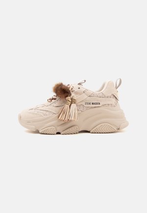 Baskets épaisses beiges en tissu texturé, avec accessoires de lacets pompons et en fourrure, et semelle épaisse rainurée, portant la marque Steve Madden sur le côté.