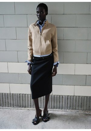 Donkere vrouw die tegen een betegelde muur staat, gekleed in een beige jack met ritssluiting, een blauw geruit overhemd, een zwarte midirok en zwarte sandalen met hak.