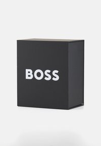 Boîte en carton noir avec une finition mate, arborant de grandes lettres blanches écrivant "BOSS" à l'avant, de forme carrée avec un design épuré.