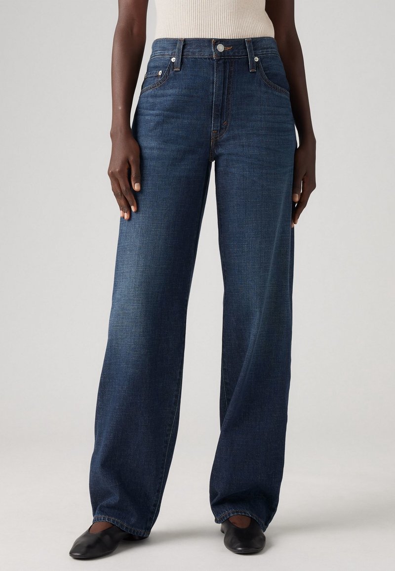 Personne portant un jean large bleu foncé, un haut beige sans manches et des chaussures plates noires, debout devant un fond clair uni.