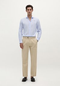 Lichtblauwe overhemden met knopen, beige broek en zwarte instappers. Het overhemd heeft een klein logo; de broek heeft voorplooien en een riemlus.
