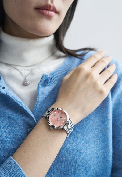 Ρολόι από ανοξείδωτο χάλυβα σε ασημί χρώμα με ροζ καντράν, φωτεινούς δείκτες και περιστρεφόμενη στεφάνη, φορεμένο σε χέρι πάνω από ένα μπλε πουλόβερ.