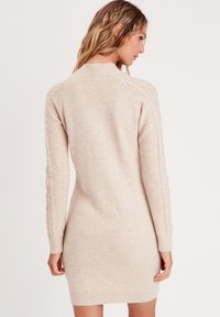 Robe pull en laine beige, avec des poignets et un ourlet côtelés. Détails en tricot torsadé le long des manches. Texture douce, longueur moyenne.