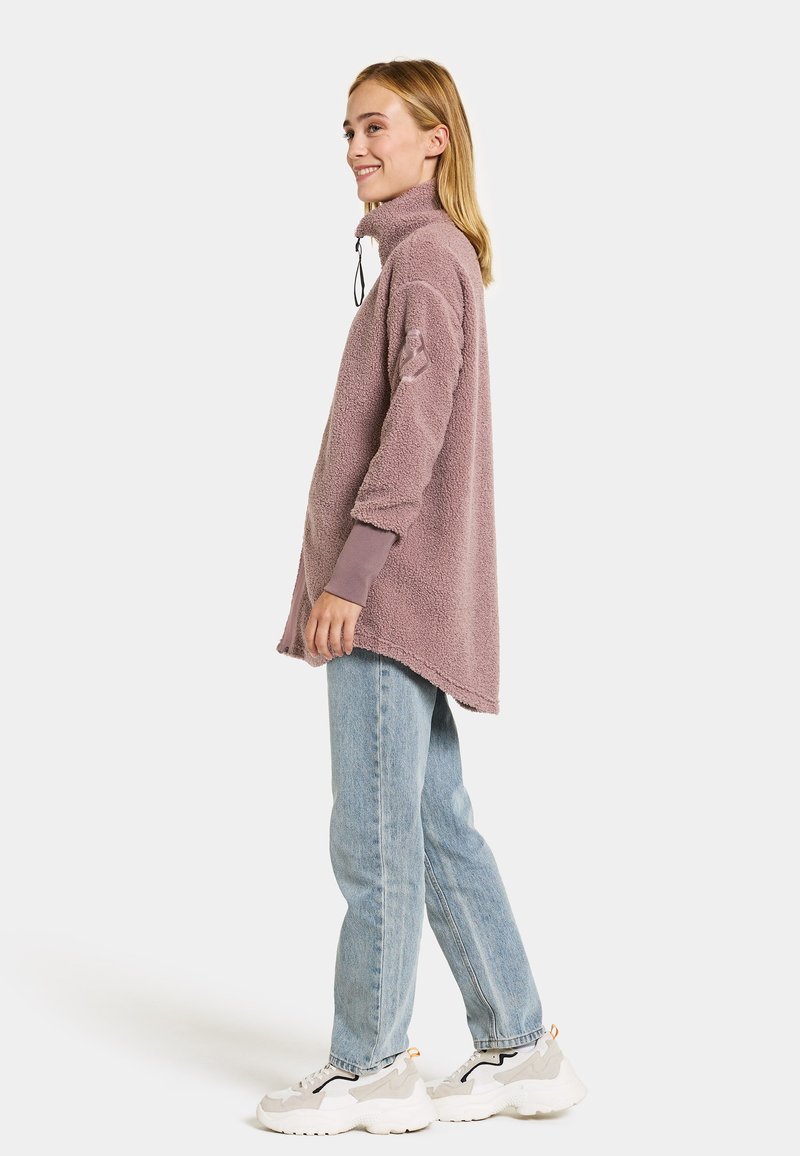 Übergroßer mauvefarbenen Sherpa-Pullover mit hohem Kragen, seitlichen Schlitzen und langen Ärmeln, kombiniert mit hellblauen, geraden Jeans und weißen Sneakers.