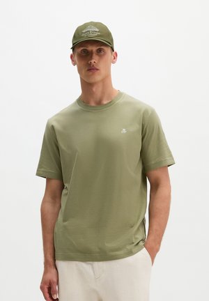 Jonge man met een olijfgroene Marc O'Polo-pet en bijpassend olijfgroen T-shirt met klein wit logo op de borst, beige broek, staand in een neutrale houding.