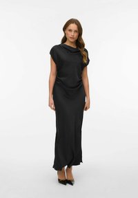 Černé šaty bez rukávů s volně splývajícím výstřihem, rozparky na kolena po stranách a hladkým materiálem, doplněné o černé lodičky s špičatou špičkou.