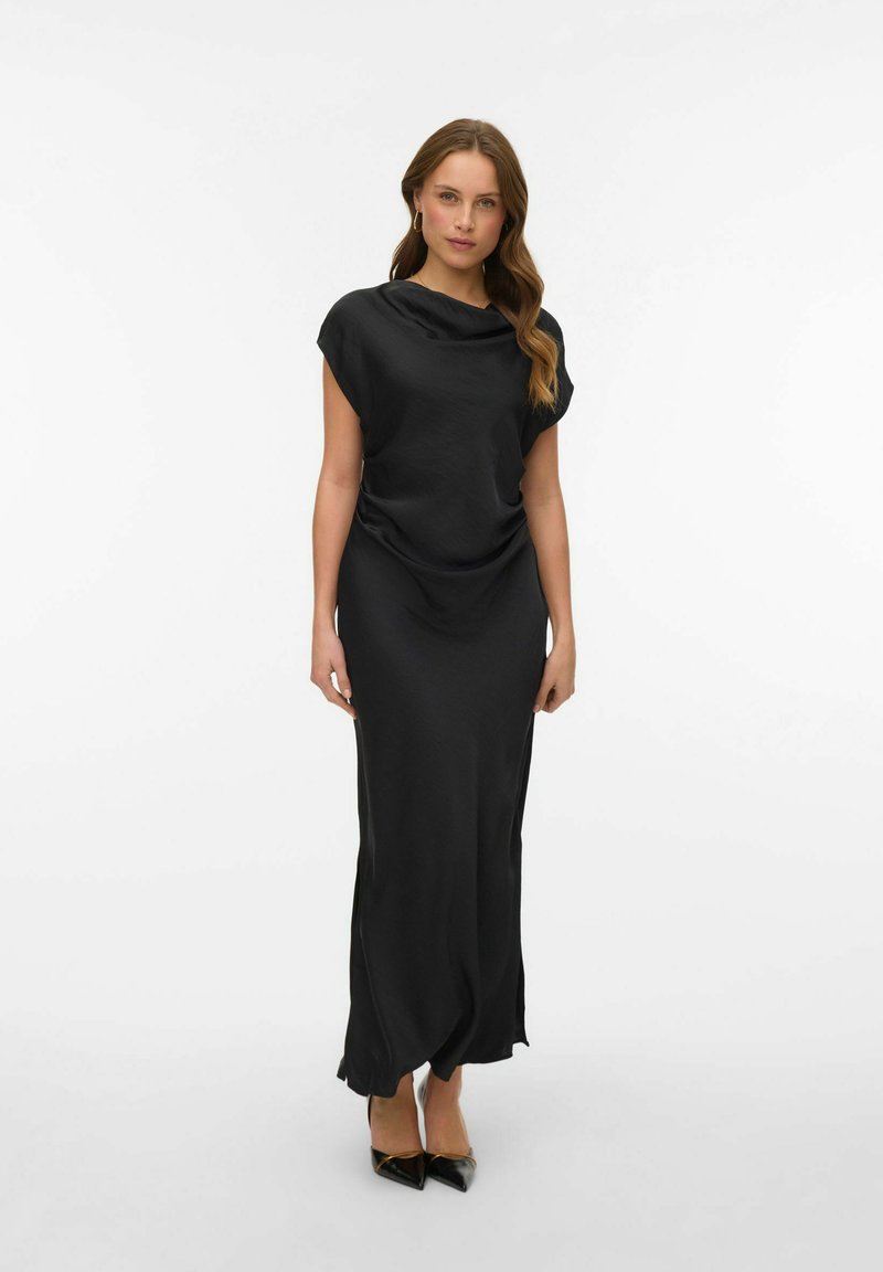 Černé šaty bez rukávů s volně splývajícím výstřihem, rozparky na kolena po stranách a hladkým materiálem, doplněné o černé lodičky s špičatou špičkou.