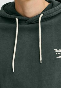Felpa con cappuccio di un verde scuro con una consistenza morbida, caratterizzata da lacci bianchi e un logo sul petto. Il design include un cappuccio spazioso.