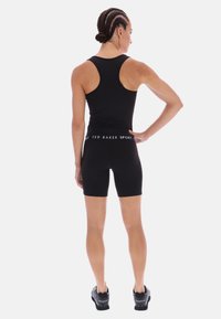 Fekete sportos ujjatlan felső versenyhát kialakítással, fekete, magas derekú rövidnadrággal párosítva, amelyen fehér logós pánt található. A modellen fekete sportcipők vannak.