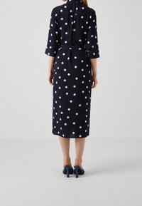 Donna in piedi con la schiena rivolta verso la fotocamera, indossa un abito midi blu navy con pois bianchi e scarpe col tacco blu.
