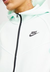 Light mint green hoodie made of soft fabric with a black zipper and Nike logo on the chest. Features a high collar and textured detailing.