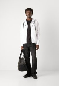 Haut à zip blanc avec cordons de serrage, t-shirt noir, jeans noirs et baskets noires. Sac gris à surface texturée tenu par le modèle.