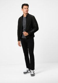 Schwarze Lederjacke mit Stehkragen, strukturierten Schulterdetails, kombiniert mit einem grauen Pullover, schwarzen Jeans und schwarz-weißen Sneakern.