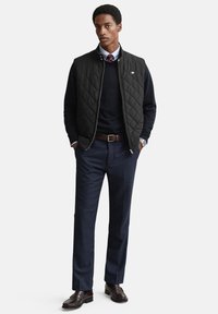 Juodas dygsniuotas liemenė virš tamsiai mėlyno megztinio, derinama su tamsiomis siūtomis kelnėmis ir rudos odos batais, turinti klasikinius apykaklę ir sagų detales.