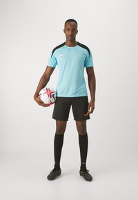 Camisa deportiva azul claro con detalles negros, pantalones cortos negros, medias hasta la rodilla negras y botas de fútbol negras. Sosteniendo un balón de fútbol con un diseño colorido.
