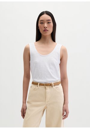 Donna con capelli lunghi e scuri che indossa un top bianco senza maniche infilato in pantaloni beige con cintura beige, in piedi davanti a uno sfondo chiaro e semplice.