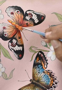 Lederen oppervlakte met roze achtergrond met geschilderde vlinders in oranje, blauw en zwart, vergezeld van bloemaccenten. Een hand houdt een kwast vast.
