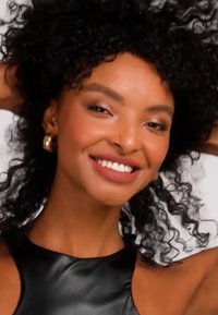 Leende kvinna med lockigt svart hår som bär guldfärgade hoops och ett ärmlöst svart linne, poserande med armarna upplyfta bakom huvudet.