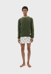 Grüner Pullover mit rundem Ausschnitt, getragen über weißen Shorts mit blauen Punkten. Das Model hat kurze, lockige Haare und ist barfuß.
