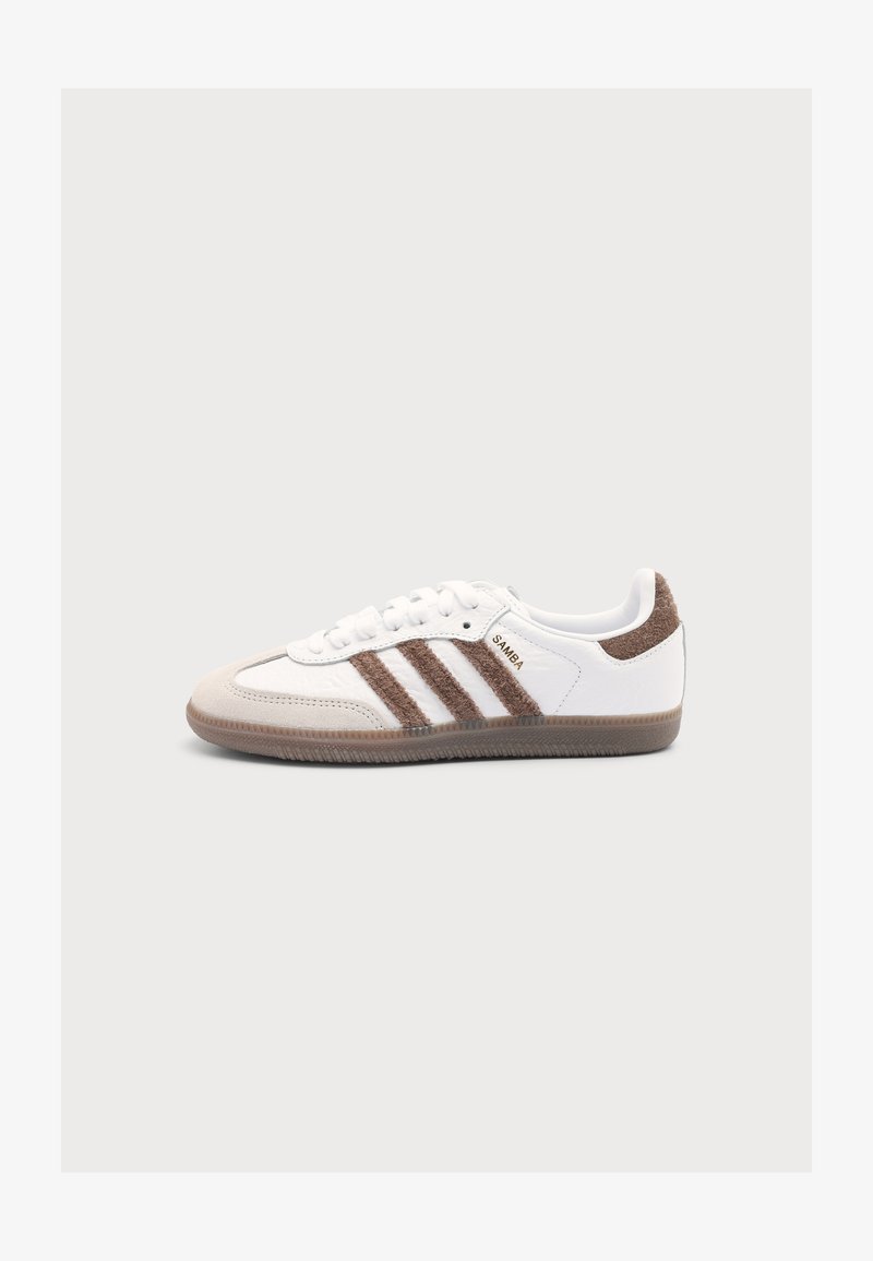 Λευκό αθλητικό παπούτσι Adidas Samba με καφέ σουέτ ρίγες, καφέ τακούνι, μπεζ κάλυμμα δαχτύλων και διάφανη λαστιχένια σόλα, εμφανίζεται από πλευράς.