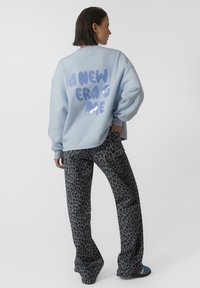 Jasnoniebieski, oversize bluza dresowa z nadrukowanym tekstem na plecach; zestawiona z szarymi spodniami w panterkę o poszerzanym fasonie. Gładki materiał z luźnym krojem.