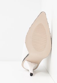 Semelle beige d'une chaussure à talon aiguilles à bout pointu avec talon stiletto blanc et petits clous métalliques le long du bord supérieur.