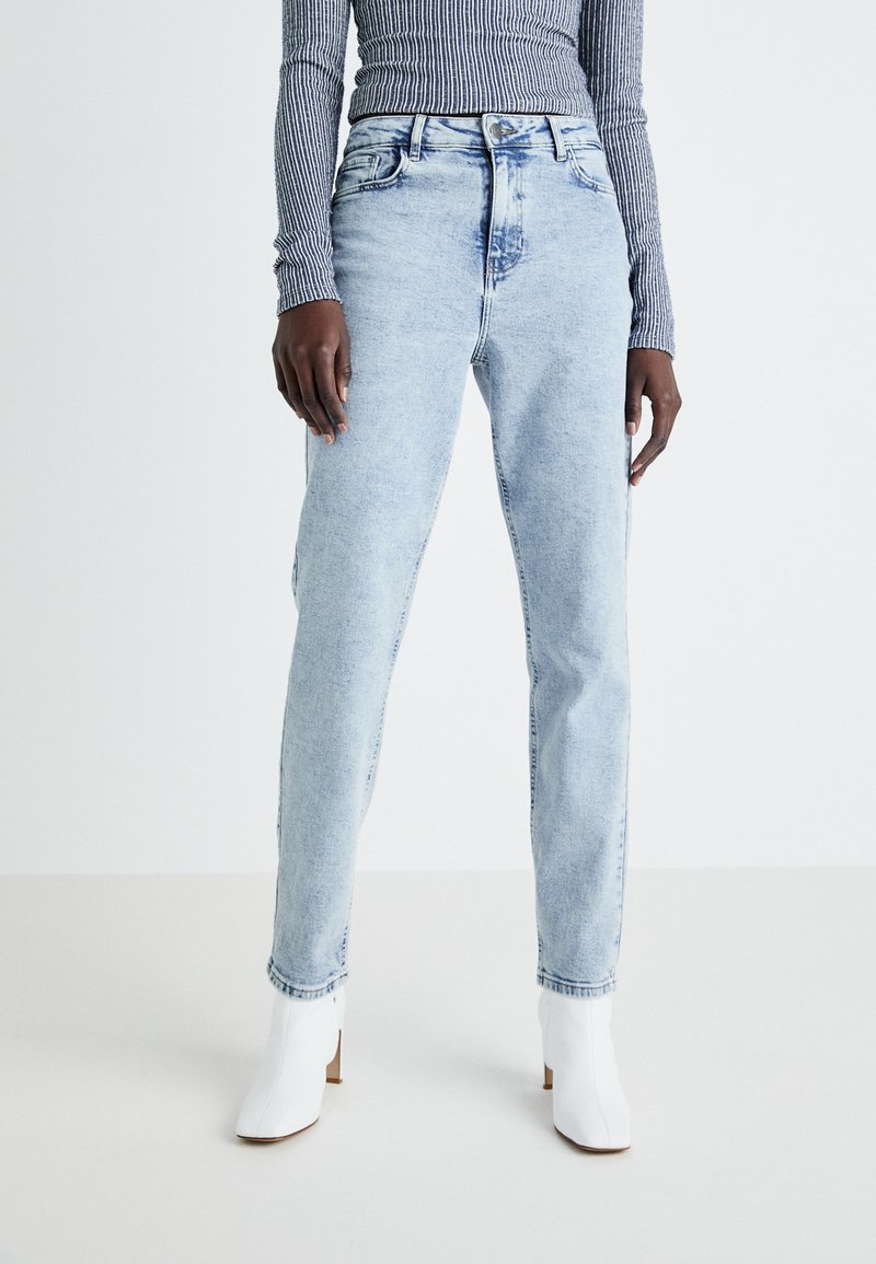 Jeans de cintura alta, azul clara, com um acabamento desbotado, apresentando cinco bolsos e um design de perna reta, combinados com botas de salto branco até o tornozelo.