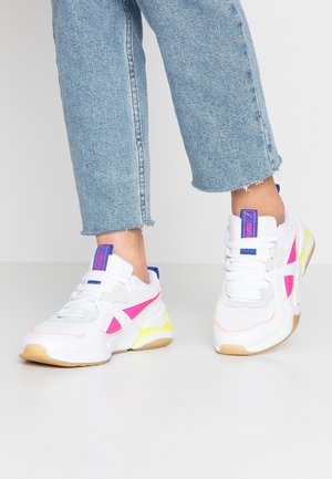 Person trägt weiße Sneakers mit pinken und gelben Akzenten und blauen Logo-Tags, kombiniert mit hellblauen, gekürzten Jeans.