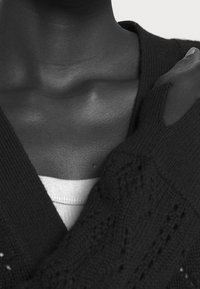 Gros plan sur le cou et l'épaule d'une personne portant un pull noir texturé sur une chemise blanche, avec une main reposant sur l'épaule.