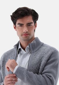 Junger Mann passt den Bund eines grauen, strukturierten Sweatshirts mit Reißverschluss über einem hellen Hemd an und schaut mit neutralem Ausdruck nach vorne.