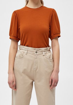 Vrouw draagt een roestbruine geribbelde top met korte mouwen en een beige broek met hoge taille en een geplooide tailleband, staand tegen een effen achtergrond.