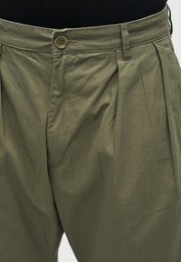 Παντελόνια λαδί με πιέτες, κούμπωμα με κουμπί, θηλιές για ζώνη και φερμουάρ, φορεμένα με μαύρο τοπ που είναι χωμένο μέσα.