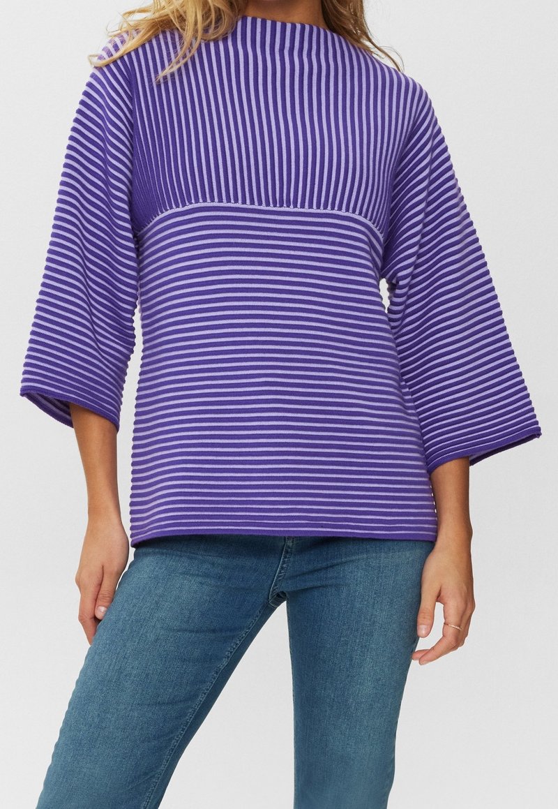 Fialový pruhovaný top s vysokým výstřihem a širokými 3/4 rukávy. Látka má žebrovanou strukturu. Doplněno modrými džínami pro kompletní vzhled.
