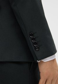 Manica di giacca verde scuro con quattro bottoni neri e una sottile trama. Polsino della camicia bianca visibile sotto la giacca.
