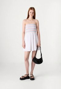 Baltos spalvos suknelė be petnešėlių, su daugiapoliu ruošiniu ir sugriebiama sijonu, derinama su juoda rankine per petį ir platformos basutėmis.