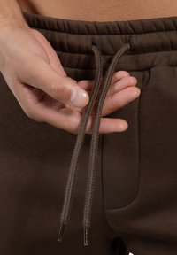Brune atletiske shorts med snøresløjfe. Stoffet er glat, og snorene er flet med plastspidser. Synlig sidelomme.
