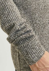 Grijze en beige gebreide trui-sleeve met gestructureerde ribbels en een geplooide manchet, met een mix van lichte en donkere garens in een warme tint.