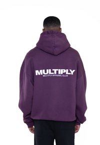 Fialová prešívaná mikina s kapucňou. Na zadnej strane sa nachádza výrazný biely text "MULTIPLY" a "MULTIPLY APPAREL CLUB." Mäkká tkanina.