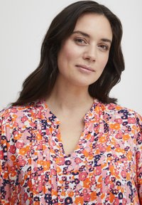 Bluză florală cu modele roz, portocalii și violet, cu decolteu în V și pliuri, realizată dintr-un material ușor.