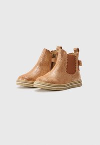 Botines Chelsea en rosa dorado brillante con bordes ondulados, paneles laterales elásticos y tiradores. Presentan una superficie texturizada y una suela de color claro.