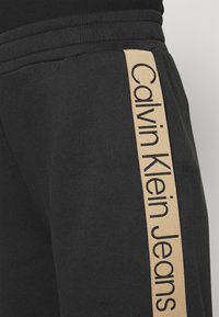 Černé teplákové kalhoty s měkkou texturou, na boku se svislým béžovým pruhem s logem "Calvin Klein Jeans".