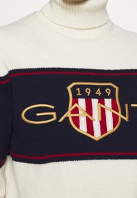 Crème col roulé trui met een marineblauwe streep en rode accenten, geborduurd 'GANT' logo en een schildontwerp in goud en rood.