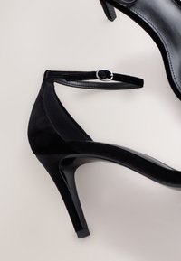 Crne cipele na visoku petu s elegantnim dizajnom, izrađene od materijala nalik antilopu s sjajnim detaljima, pričvršćene nježnim remenom oko gležnja.