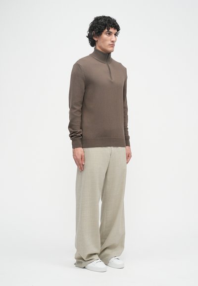Pull en maille marron à manches longues avec un col montant et une demi-fermeture éclair, associé à un pantalon large beige clair et des baskets blanches.
