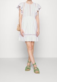 Vestido de lavanda claro con mangas con volantes, cinturilla fruncida y dobladillo con volantes. Combinado con sandalias trenzadas verdes y un pequeño bolso beige.