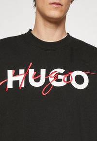 Sudadera negra con gran logo blanco "HUGO" y acentos en cursiva rojos. Cuello acanalado y textura típica de la tela de algodón.