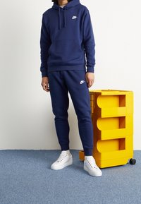 Marinblå Nike-hoodie och joggers med en vit logotyp, matchat med vita sneakers. Står bredvid en lysande gul förvaringsvagn på blå matta.